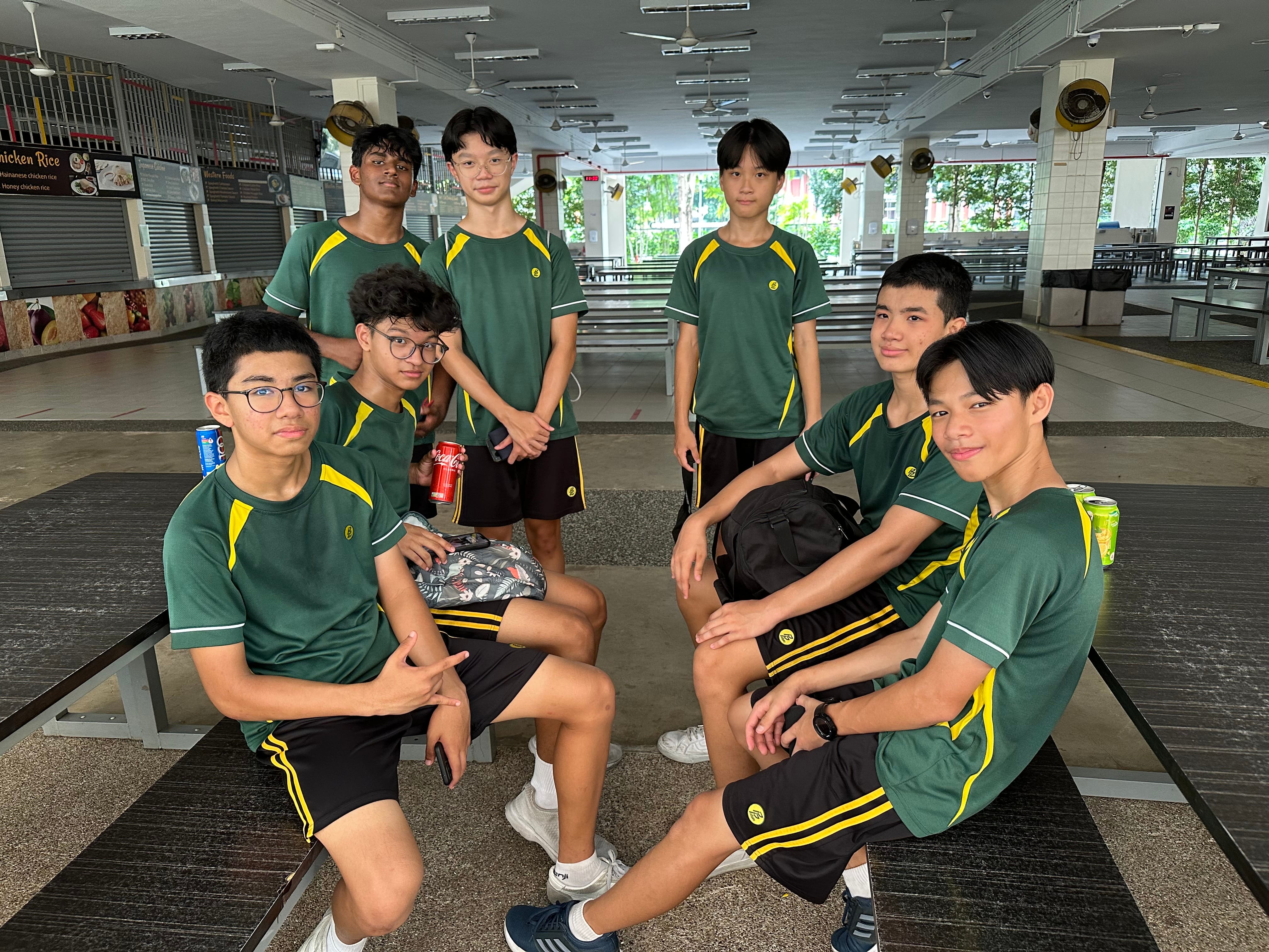 Seven students in green sports uniforms pose on cafeteria benches, some seated, some standing.